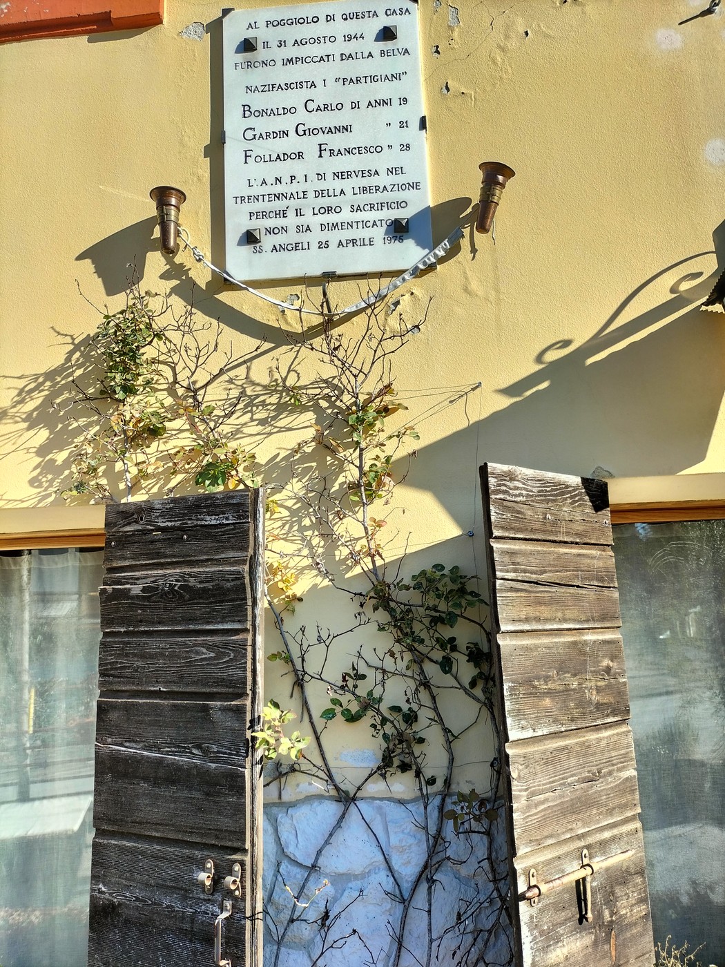 Lapide commemorativa a Bonaldo, Gardin e Follador su una parete con porte in legno e vegetazione circostante.
