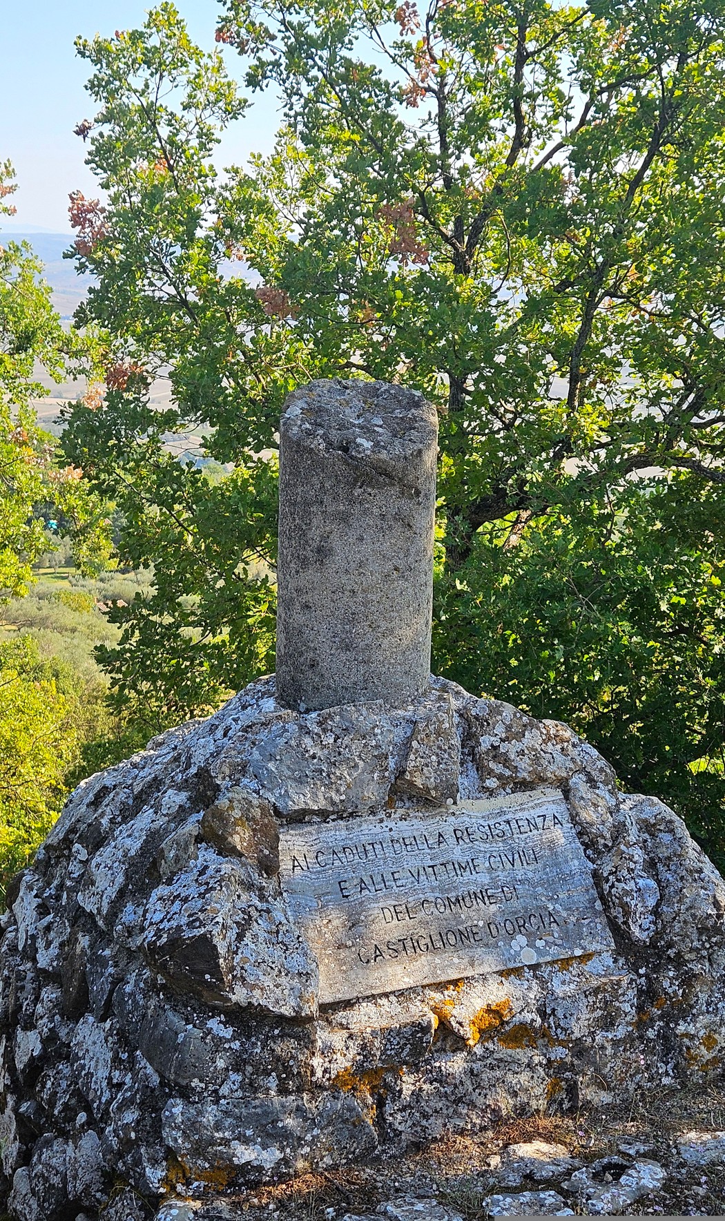 Cippo in pietra con colonnina sommitale e lapide incisa, tra gli alberi di un rilievo collinare.