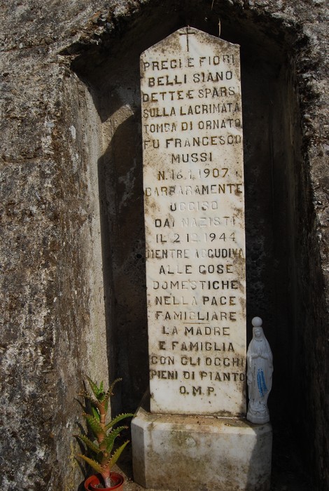 Cippo a Mussi con iscrizione commemorativa, situato in una nicchia, circondato da piante e una statua.
