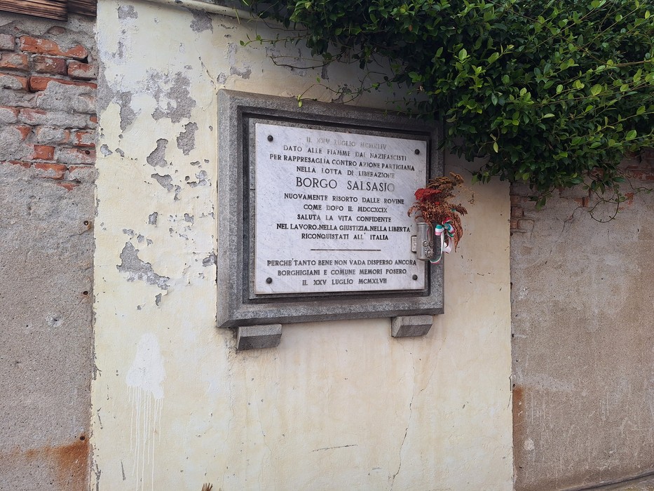 Lapide in marmo con iscrizione, fissata su muro esterno intonacato, con un piccolo fiore decorativo appeso accanto.