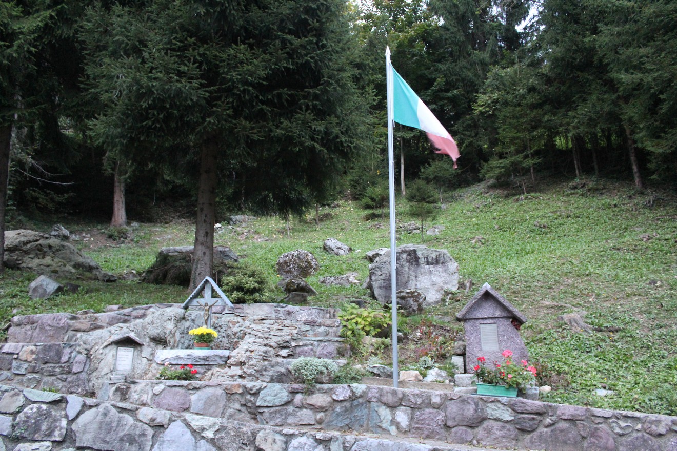 Monumento commemorativo con bandiera italiana e paesaggio naturale circostante.