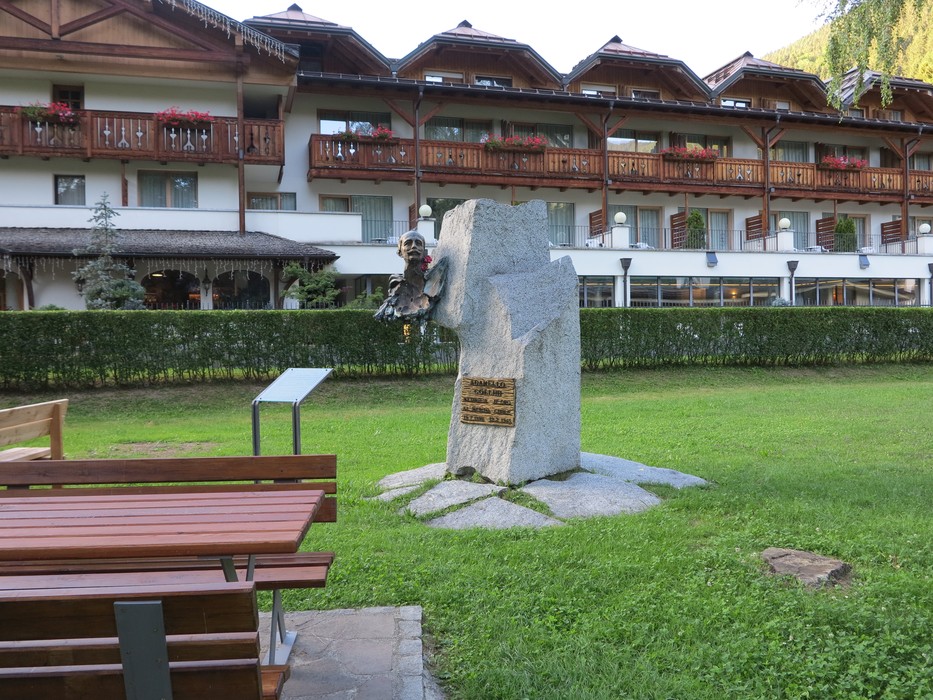 Monumento a Collini in uno spazio verde, circondato da panchine e un edificio montano.
