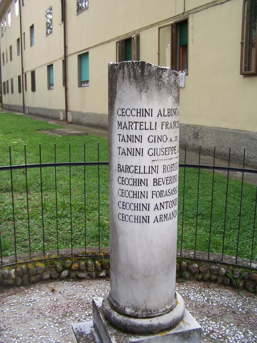 Cippo commemorativo con nomi incisi, situato in un'area verde circondata da una balaustra.