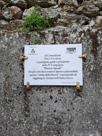 Targa bianca con iscrizione, fissata su muro in pietra con staffe dorate, tra erba e rocce.