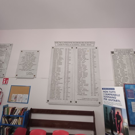 Parete con lapidi ai caduti recanti lunghi elenchi di nomi, in una sala con libreria e sedie rosse.