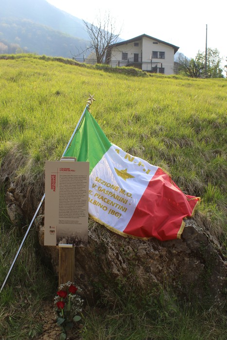 Impronta partigiana con bandiera italiana e targa commemorativa in un paesaggio montano.