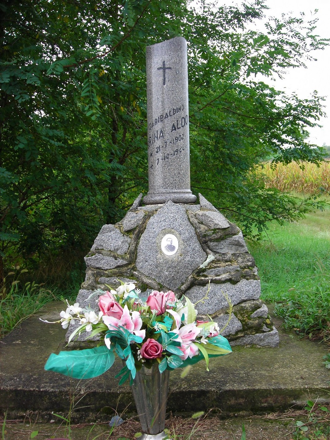 Cippo commemorativo con fiori artificiali, situato in un'area verde. Rappresenta un luogo di memoria e rispetto.