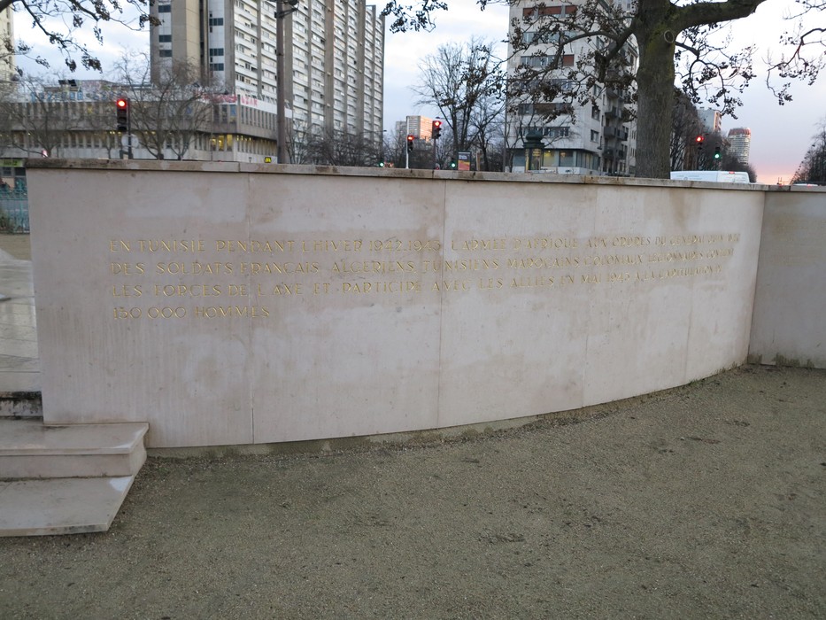 Statua a Juin in un contesto urbano, con un murale che riporta un testo inciso su pietra.