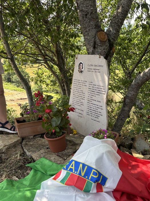 Lapide commemorativa di Giacomini, con fiori e bandiere, immersa in un paesaggio naturale.