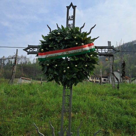 Croce a Volpato adornata da un corona di fiori, in un paesaggio naturale con vigna sullo sfondo.
