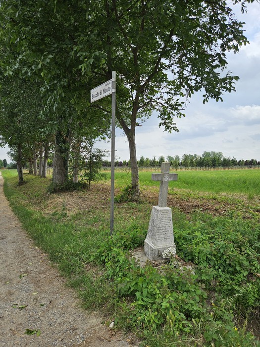 Cippo in pietra con croce, circondato da alberi e campo verde, lungo una strada sterrata.