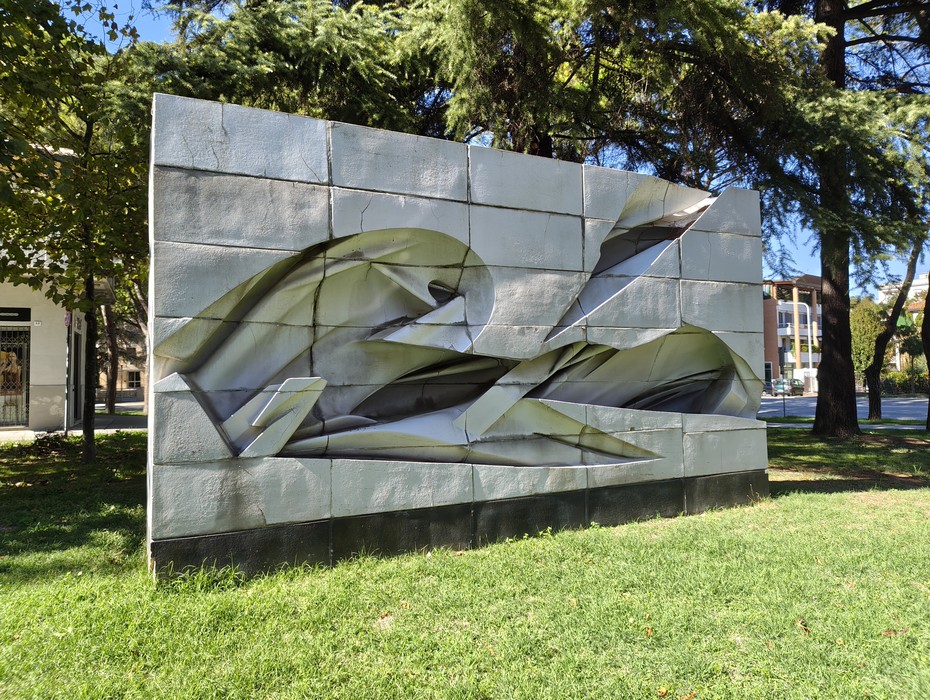 Monumento alla Resistenza in pietra grigia con figure astratte in rilievo, collocato su un prato tra gli alberi.