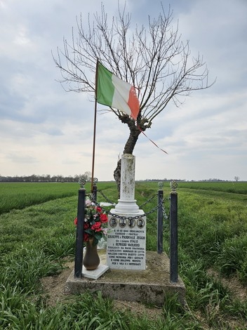 Cippo commemorativo con bandiera italiana, circondato da fiori, in un paesaggio verdeggiante.