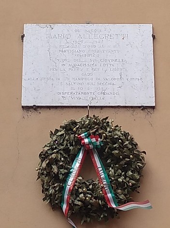 Lapide commemorativa di Mario Allegretti con un'omaggio floreale e un nastro tricolore.
