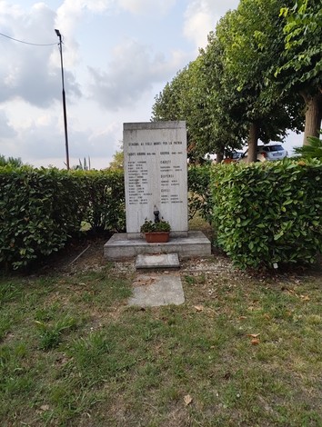 Monumento in pietra con iscrizione, circondato da siepi, in un'area verde con un vaso di fiori davanti.