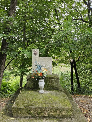 Cippo a Ghirardi circondato da vegetazione, con decorazioni floreali e un vaso in pietra.