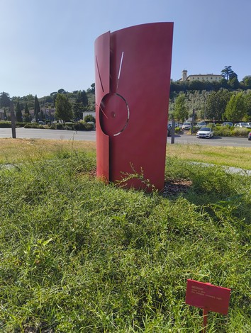 Scultura rossa in un'area verde, simbolo di resistenza femminile, con sfondo di un paesaggio collinare.