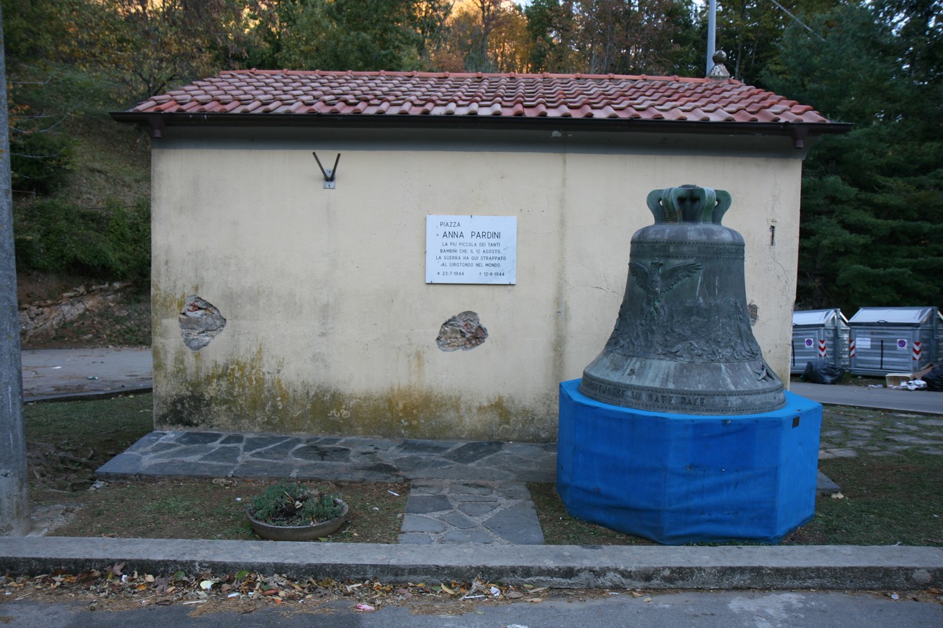 Lapide commemorativa di Annal Pardini con una campana all'esterno, situata in un contesto naturale.