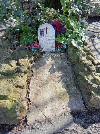 Rappresentazione del cippo commemorativo di Bruno Baldinotti e Carlo Lazzerini, adornato da fiori e vegetazione.