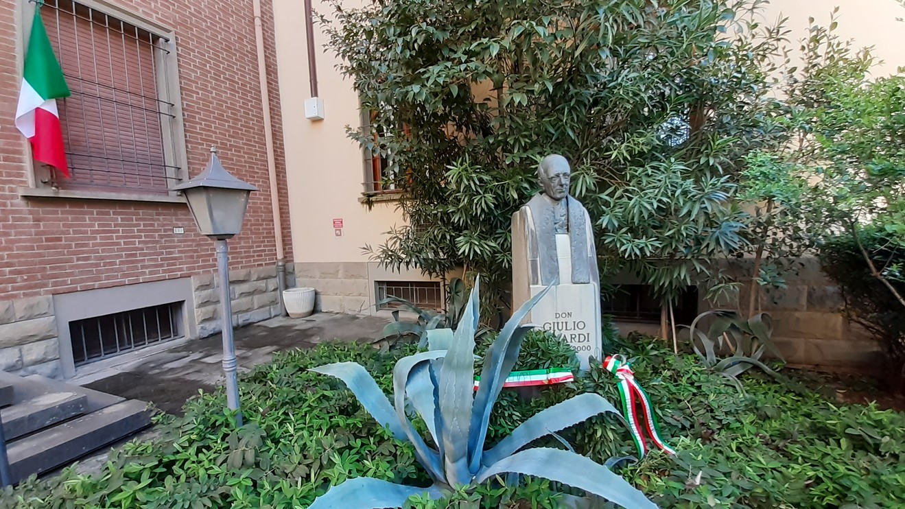 Statua commemorativa di don Minardi, circondata da piante in un cortile con bandiera italiana.