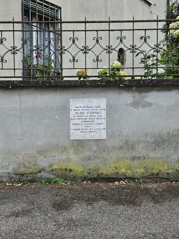 Lapide commemorativa con iscrizioni su un muro, circondata da vegetazione e ferro battuto.