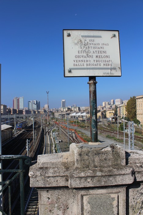Lapide commemorativa che ricorda Atzeni e Meloni, con vista sui binari ferroviari e il panorama urbano sullo sfondo.