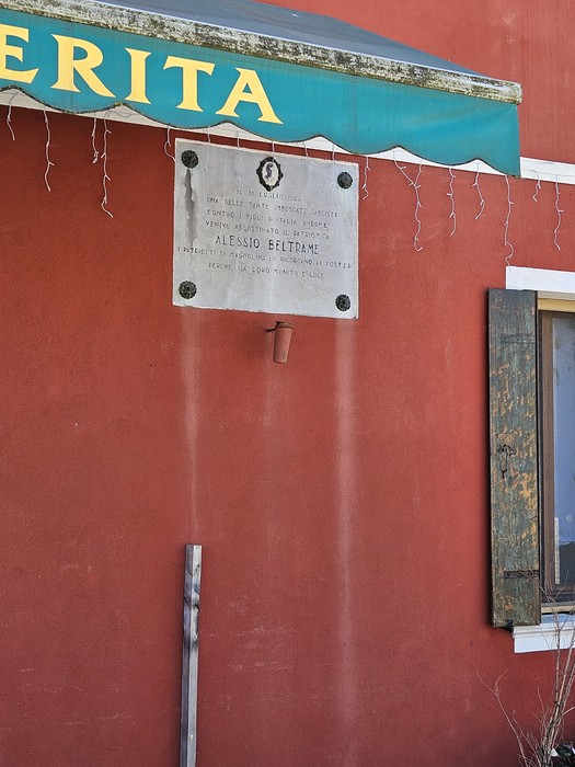 Lapide commemorativa su un muro rosso, con tettoia verde e decorazioni, dedicata a Alessio Beltrame.
