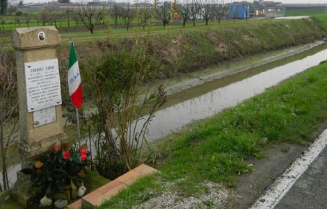Cippo commemorativo a Savioli, con bandiera italiana e fiori, lungo un canale in un paesaggio rurale.