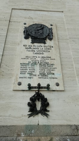Lapide in marmo con iscrizione e bassorilievo, affissa a una parete, con corona in metallo sottostante.