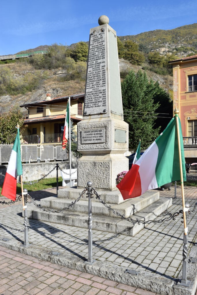 Obelisco in pietra con iscrizione, su basamento in piazza, circondato da bandiere italiane e catene.