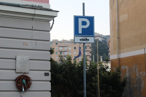 Lapide commemorativa accanto a un cartello stradale di sosta, con edifici sullo sfondo.
