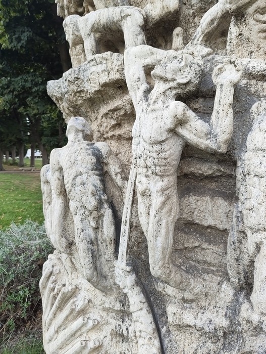 Monumento in pietra con figure umane scolpite in rilievo, collocato all’aperto in un’area alberata.
