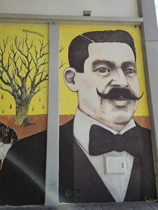 Murale colorato che ritrae un uomo con baffi, indossando un tailleur, sullo sfondo di un albero spoglio.