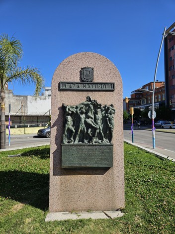 Monumento a Matteotti, in pietra con rilievi bronzei, circondato da vegetazione e con cielo azzurro sullo sfondo.