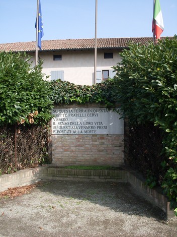 Lapide commemorativa dedicata ai sette frati cervi, circondata da vegetazione e bandiere.