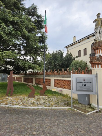 Largo Martiri del Nazifascismo, con statue e bandiera italiana tra alberi e muri storici.