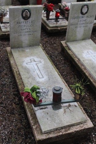 Tombe di pietra con croce, candele e fiori, dedicate ai caduti per la Libertà. Atmosfera solenne e rispettosa.