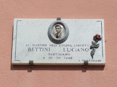 Lapide commemorativa dedicata a Luciano Bettini, partigiano, con foto e decorazioni floreali.