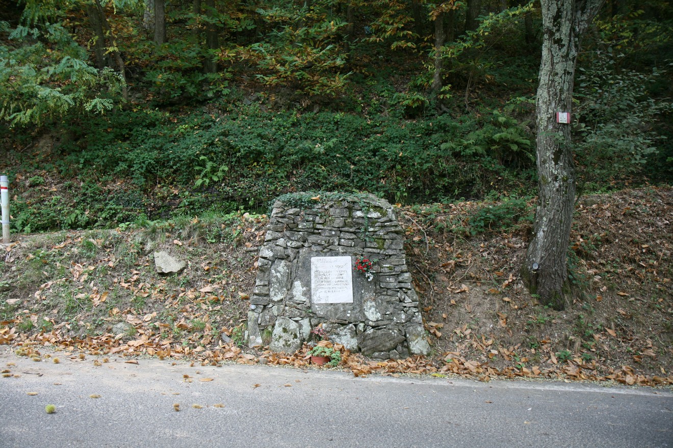 Lapide commemorativa a Ighenad e Mugik, immersa nella natura, lungo una strada nel bosco.
