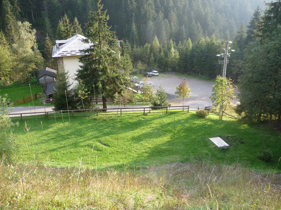 Monumento ai Dodici Caduti immerso nella natura, circondato da alberi e con vista su un parcheggio e un edificio.