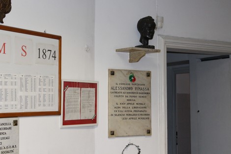 Lapide commemorativa dedicata ad Alessandro Rimassa, situata in un ambiente interno con opere murali.