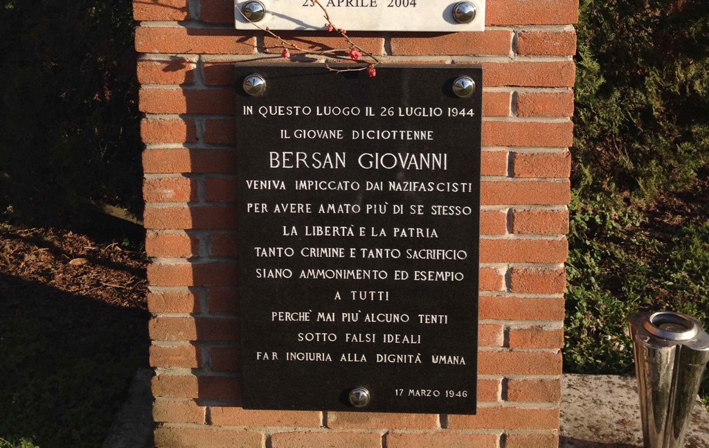Targa in marmo nero con iscrizione, fissata a una parete in mattoni, con fiori appesi sopra.