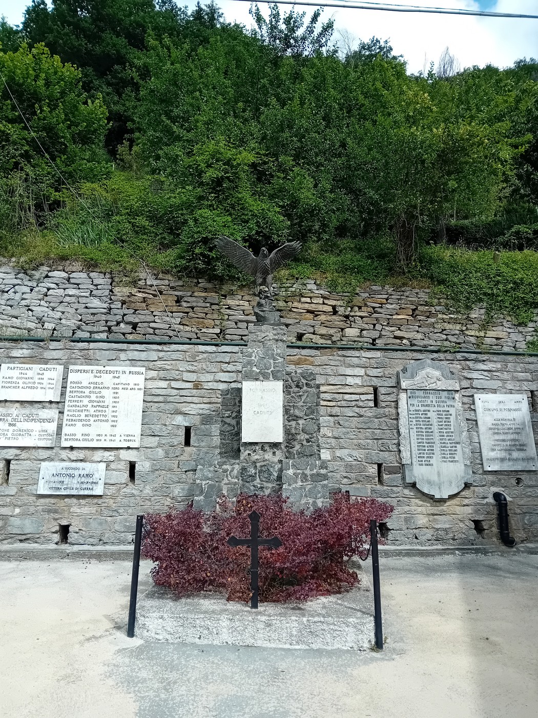 Monumento ai caduti circondato da pareti di pietra e vegetazione, con iscrizioni memoriali sui lati.