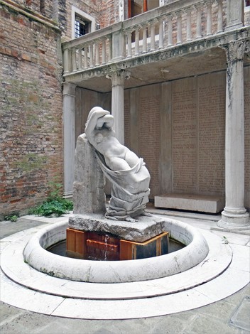 Scultura di un giovane che emerge da un basamento, circondata da un ambiente monumentale e storico.
