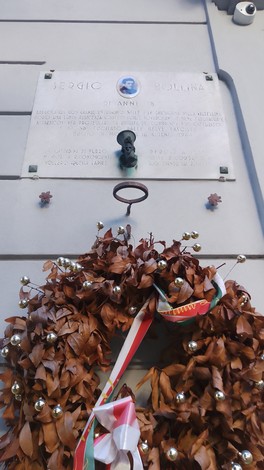 Lapide commemorativa di Sergio Bollina, adornata da una ghirlanda di foglie e nastri colorati.