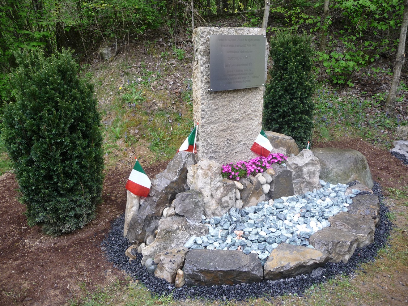 Cippo commemorativo ad Adobati, circondato da pietre, fiori e bandiere italiane, immerso nella natura.