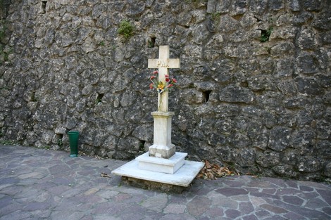 Croce commemorativa per le vittime di Forno, adornata con fiori, contro un muro in pietra.