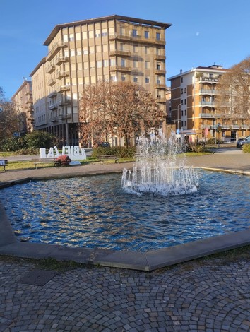 Fontana circolare con getti d'acqua, situata in un'area verde urbana, circondata da edifici e alberi.