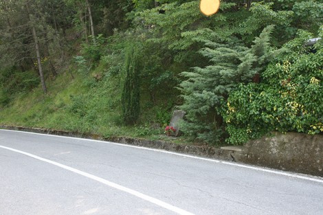 Cippo commemorativo immerso nella vegetazione lungo una strada tortuosa.