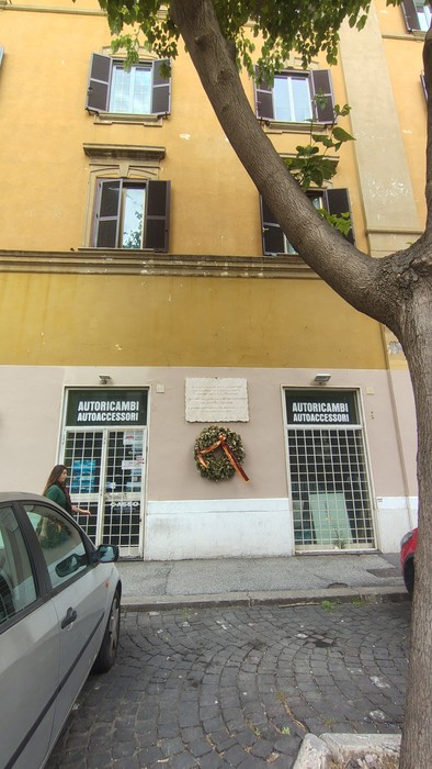 Lapide commemorativa su un edificio a Gallarello, con decorazione natalizia e negozi di ricambi auto in evidenza.